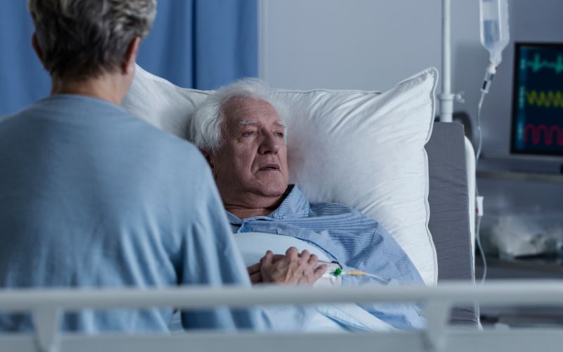 Bliska osoba trzyma za rękę Seniora z zaawansowaną demencją leżącego w łóżku szpitalnym